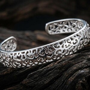 May include: A silver-toned cuff bracelet featuring an intricate, openwork filigree pattern that covers the entire outer surface. The bracelet has a C-shaped design with a wide opening, showcasing a polished metallic finish and detailed scrollwork.