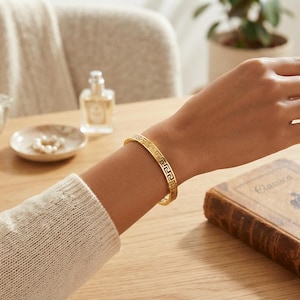 May include: A gold-toned metal bangle bracelet featuring a continuous cut-out Greek key pattern. The bracelet has a polished finish and a rigid, circular structure that encircles the wrist, showcasing a repeating geometric design throughout.