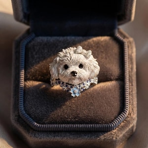 May include: A silver-toned ring featuring a sculpted dog head with a small bow on its head. The band is adorned with small blue and clear stones, and a single light blue flower charm hangs from the center of the dog's neck area.