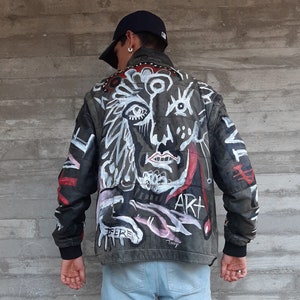 Puede incluir: Una chaqueta de cuero negra con un dise&ntilde;o pintado a mano en la espalda. El dise&ntilde;o presenta una cara abstracta blanca y negra con acentos rojos y rosados. El texto "ART" es visible en la parte posterior de la chaqueta.