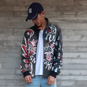 Puede incluir: Una chaqueta de cuero negra con un dise&ntilde;o abstracto blanco, rojo y negro. La chaqueta tiene una cremallera y un cuello alto. La persona que lleva la chaqueta tambi&eacute;n lleva una camiseta blanca y unos vaqueros azules.