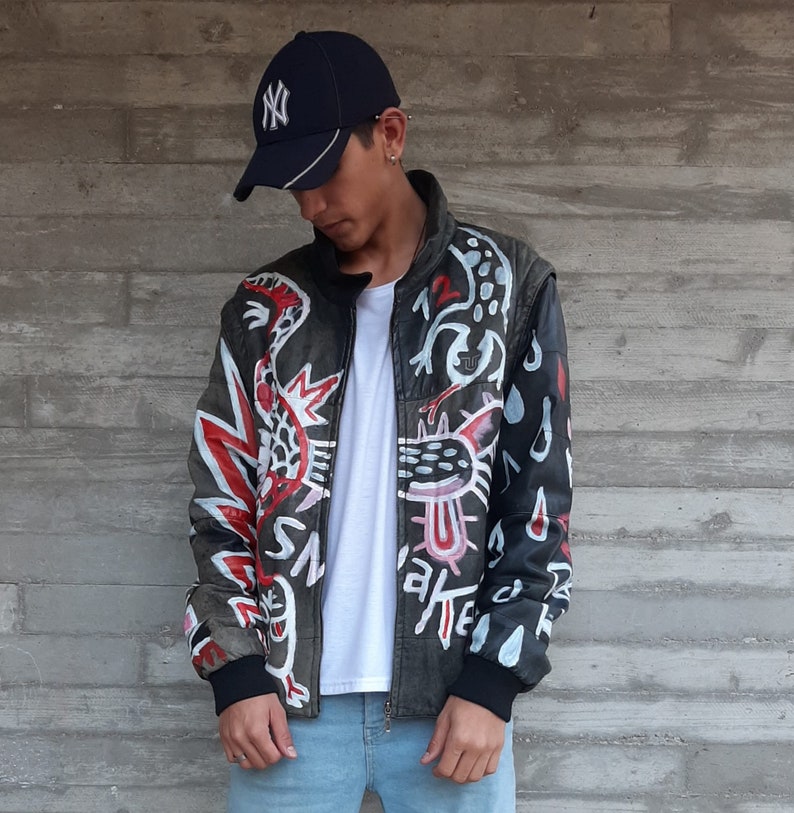 Puede incluir: Una chaqueta de cuero negra con un dise&ntilde;o abstracto blanco, rojo y negro. La chaqueta tiene una cremallera y un cuello alto. La persona que lleva la chaqueta tambi&eacute;n lleva una camiseta blanca y unos vaqueros azules.
