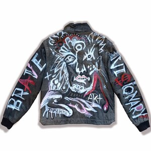 Puede incluir: Chaqueta de cuero negra con un dise&ntilde;o pintado a mano que presenta una cara, formas abstractas y las palabras "Brave", "Art", "Different" y "Revolutionary".