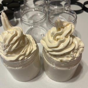 Può includere: Due piccoli vasetti di plastica trasparente riempiti di crema bianca, sormontati da un motivo a spirale. Vasetti vuoti e coperchi neri sullo sfondo suggeriscono un prodotto cosmetico.