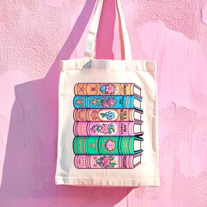 Op de afbeelding: Een witte canvas tote bag met een kleurrijke illustratie van zes boeken die op elkaar gestapeld zijn. De boeken hebben bloemmotieven en de titels zijn "Pride & Prejudice", "Mansfield Park", "Emma", "Persuasion", "Sense & Sensibility" en "Jane Austen".