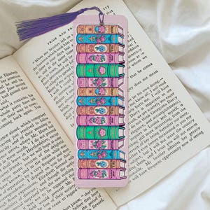 May include: A bookmark with a design of stacked books, each adorned with floral patterns. The bookmark has a purple tassel and is placed on an open book. The books are in pink, green, and blue hues.