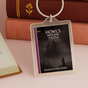 May include: A rectangular keyring displaying the cover of "Howl's Moving Castle" by Diana Wynne Jones. The artwork depicts a dark castle, a figure, and the book's title. The keyring has a silver-coloured ring and a clear plastic frame.