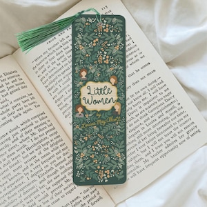 May include: A bookmark featuring a floral design in green and cream, with the title "Little Women" by Louisa May Alcott. It has a green tassel and lies on an open book.