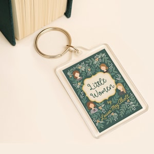 Könnte beinhalten: Ein rechteckiger Schlüsselanhänger mit dem Buchtitel "Little Women" von Louisa May Alcott. Das Design umfasst Illustrationen von Frauen und florale Elemente auf einem dunkelgrünen Hintergrund. Der Schlüsselanhänger hat einen transparenten Kunststoffrand und einen silbernen Schlüsselring.