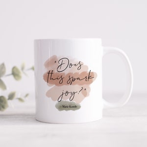 May include: White ceramic mug with a quote from Marie Kondo: "Does this spark joy?" The quote is written in black cursive script on a background of watercolor-style brown and beige shapes.