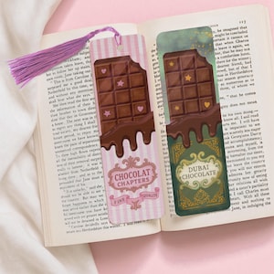 Op de afbeelding: Twee decoratieve bladwijzers met chocoladereepontwerpen. Eén is roze met de tekst "Chocolat Chapters", de andere is groen met de tekst "Dubai Chocolate". Beide hebben een chocolade-druppeleffect en ster- en hartaccenten.