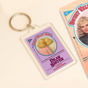 May include: A clear acrylic keychain featuring a rectangular insert with the text "Sweet Valley High" and "Dear Sister" by Francine Pascal. The insert has an illustration of two blonde-haired figures. A book with the same theme is visible.
