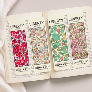 Op de afbeelding: Vier rechthoekige bladwijzers met bloempatronen en de tekst "LIBERTY LONDON" en "LIBERTY & CO. LTD. LONDON EST.1875". De bladwijzers hebben rode, blauwe, groene en roze bloemmotieven. Ze liggen op een open boek.