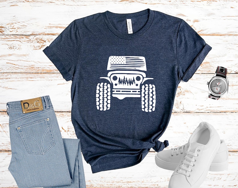 Puede incluir: Una camiseta azul marino con un gr&aacute;fico blanco de un Jeep con una bandera estadounidense en el cap&oacute;. El Jeep est&aacute; de frente y tiene neum&aacute;ticos grandes. La camiseta est&aacute; colocada sobre una superficie de madera blanca con un par de pantalones vaqueros azules, un reloj blanco y un par de zapatillas blancas.