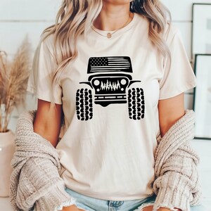 Puede incluir: Una camiseta blanca con una silueta negra de un Jeep con una bandera americana en el cap&oacute;. El Jeep tiene neum&aacute;ticos negros y una parrilla negra.