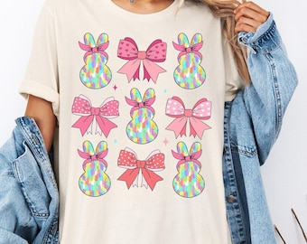 Può includere: Una maglietta beige chiaro con un design di coniglietto pasquale colorato e un fiocco. I coniglietti indossano fiocchi rosa e hanno un motivo arcobaleno.