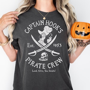 May include: Dark gray t-shirt featuring a Captain Hook's Pirate Crew graphic. The design includes an illustration of Captain Hook, crossed swords, a skull, and the text "Est. 1953". A pumpkin-shaped mug is held in the hand.