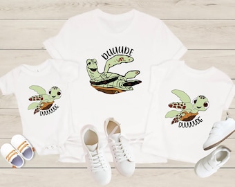May include: Three white t-shirts with a green sea turtle graphic and the text "DUDE" on each. The shirts are in different sizes and are laid out on a wooden surface. There are also three pairs of white sneakers.