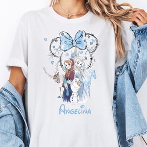 Könnte beinhalten: Weißes T-Shirt mit einem hellblauen Schleifendesign, Herzen und Sternen sowie einer Skizze von Figuren aus dem Film Frozen. Der Name "Angelina" ist in hellblauer Schrift darunter geschrieben.