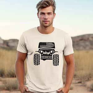 Puede incluir: Una camiseta blanca con una silueta negra de un Jeep con una bandera estadounidense en el cap&oacute;. El Jeep tiene neum&aacute;ticos grandes y una parrilla con una escena de bosque en su interior.