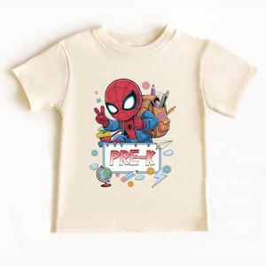 May include: Cream-colored t-shirt featuring a cartoon Spider-Man character with a backpack, pencils, and a sign that says "PRE-K." The design includes a globe, flowers, and lightning bolts. The character is giving a peace sign.