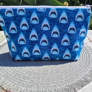 May include: A blue zippered pouch with a white zipper and a repeating pattern of white shark teeth on a blue background.