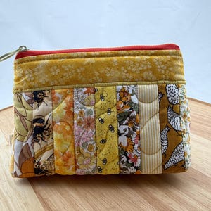May include: A quilted pouch with a zip closure. The rectangular pouch has a patchwork design with floral and insect fabrics in yellow, orange, and white. The zip is red, and the pull is green.
