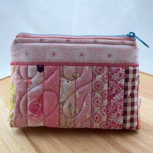May include: A quilted pouch with a light blue zip. The pouch is rectangular and made from various pink fabrics with floral and polka dot patterns. The stitching is visible, and the design is handmade.