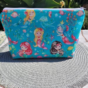 May include: A turquoise zippered pouch with a repeating pattern of mermaids, fish, and seashells. The mermaids are all wearing crowns and have long flowing hair.