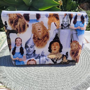 May include: White zippered pouch featuring a colourful collage of characters from The Wizard of Oz. The characters include Dorothy, the Scarecrow, the Tin Man, the Cowardly Lion, and the Wicked Witch of the West.