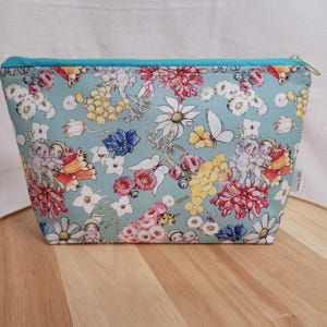May include: A turquoise zippered pouch with a floral print featuring small fairies, butterflies, and various flowers. The print is on a light blue background.