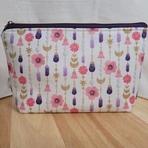 May include: A white zippered pouch with a purple zipper. The pouch has a repeating pattern of pink flowers, purple crystals, gold moons, and gold stars.
