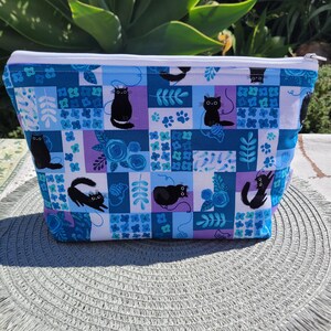 May include: A white zippered pouch with a blue and purple patchwork design featuring black cats and floral motifs.