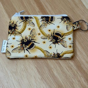 May include: A small, rectangular coin purse with a zip closure and a key ring. The purse displays a repeating pattern of bumblebees and yellow flowers on a cream background. The zip is silver, and the purse has a small tag with text.