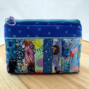 May include: A patchwork zipped pouch featuring a variety of fabric patterns in shades of blue, purple, and green. The pouch has a blue zip and a purple ring pull. The design includes floral, geometric, and character prints.