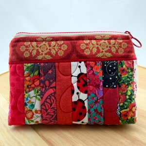 May include: A quilted fabric pouch with a red zip and pull. The pouch is made of vertical strips of patterned fabric in red, white, and black. Some strips feature floral designs and ladybirds. The top section has a red floral pattern.