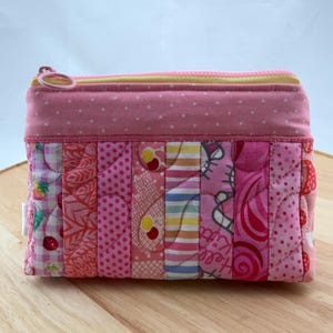 May include: A small, rectangular pink quilted pouch with a zip closure. The pouch is made of various pink patterned fabrics, including polka dots, strawberries, and a cartoon character. The zip is yellow and the pouch is on a wooden surface.