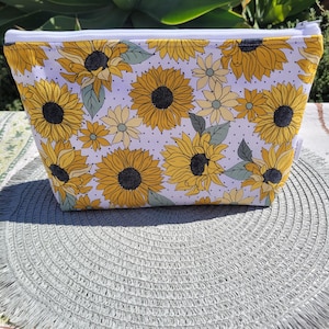May include: White zippered pouch with a sunflower print. The pouch has a white background with yellow sunflowers and small white polka dots.