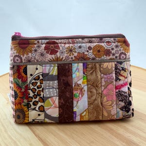 May include: A patchwork zipped pouch with diverse fabric patterns. The pouch has a floral print at the top and a mix of sunflower, leopard, and geometric designs on the front. The zip is pink, and the pouch is on a wooden surface.