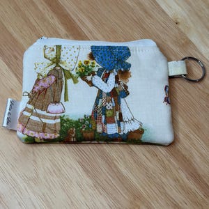 May include: A small, rectangular coin purse with a zip closure and a key ring. The purse features a vintage-style illustration of two girls in bonnets, one holding flowers. The background is a light cream colour, and the purse has the text "Lotta & Lou".