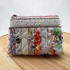 May include: A rectangular, quilted pouch with a red zip. The pouch is made of fabric strips with floral, bee, and geometric patterns. The top section has a white background with colourful triangles.