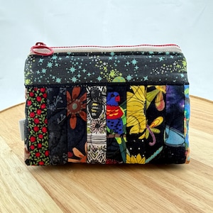 May include: A small, rectangular quilted purse with a zip closure. The purse has a patchwork design with various fabric patterns, including floral, star, and bird motifs. The zip is red, and the overall colour scheme is dark with vibrant accents.