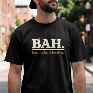 May include: A black short-sleeved t-shirt featuring the text BAH. in large white capital letters with a small red triangle above the A. Below this, the text but make it festive. is printed in smaller red and green lowercase letters.