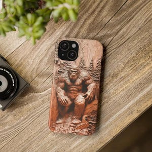 May include: A brown phone case with a carved wooden design of Bigfoot sitting on a tree stump. The case has a camera lens cutout and a textured wood grain pattern.