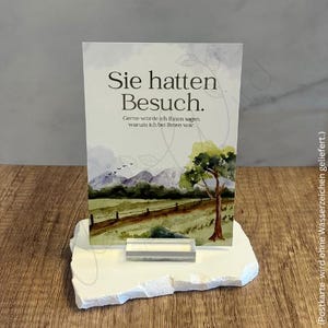 May include: A white card with the text "Sie hatten Besuch" and a watercolor landscape scene of a field, fence, and mountains. The card is displayed on a white, irregularly shaped stone base with a clear acrylic stand.