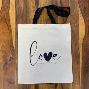 Könnte beinhalten: Eine weiße Canvas-Tragetasche mit schwarzen Henkeln und dem Aufdruck "love never fails" auf der Vorderseite. Das Wort "love" ist mit einem schwarzen Herzen in der Mitte des "o" stilisiert.