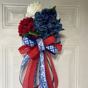 Puede incluir: Una corona de puerta patriótica con flores artificiales rojas, blancas y azules y cintas decorativas. La corona incluye un lazo grande con cintas rojas y blancas con estampado de estrellas, cintas azules con estampado floral y una cinta roja a cuadros.