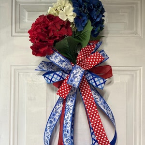 Puede incluir: Un adorno patriótico para puerta con hortensias rojas, blancas y azules y cintas decorativas. Las cintas tienen diseños de estrellas, flores y cuadros. El arreglo floral está sujeto a una puerta blanca.