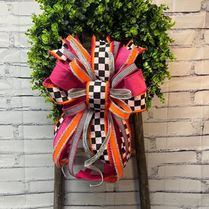 May include: A decorative wreath featuring lush green foliage adorned with a large, multi-layered bow. The bow consists of ribbons in vibrant hot pink, bright orange, and a black and white chequered pattern, accented with silver glittered edges.