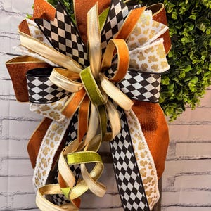 May include: A decorative bow featuring a mix of ribbons in burnt orange, black and white diamond patterns, and white with gold leopard print. The bow includes solid olive green velvet ribbon accents and long, trailing ribbon tails.